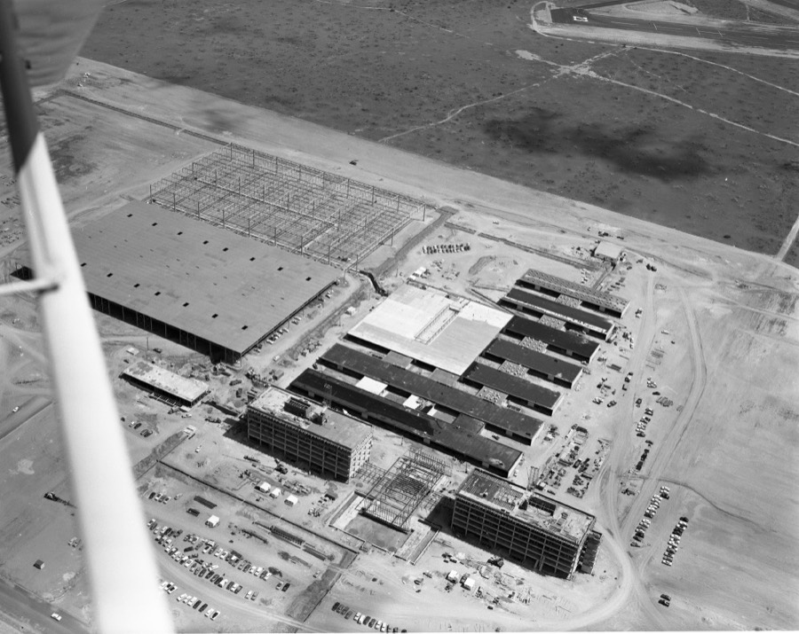 A 1957 aerial photograph. A large factory complex under construction fills the lower half of the frame, ringed by parked cars and bare graded ground. Beyond it the mesa runs to the horizon as open scrub crossed by a few dirt tracks.
